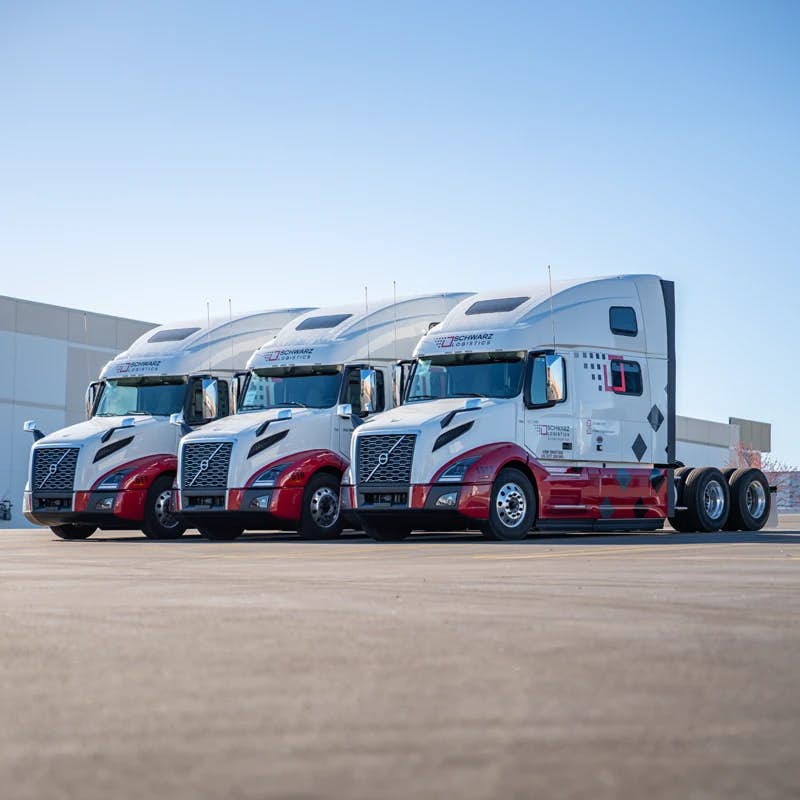 This image displays three semi-trucks in a diagonal line formation, parked on what appears to be an open, paved area. The trucks have a distinct color scheme, predominantly white with red and black graphics, and feature the Volvo logo on the front, indicating the make of the vehicles. The cabs of the trucks are large, suggesting they might be sleeper models, equipped for long-haul trips. Each truck bears the branding "SCHWARZ LOGISTICS" on the side, hinting that they are part of a commercial fleet.