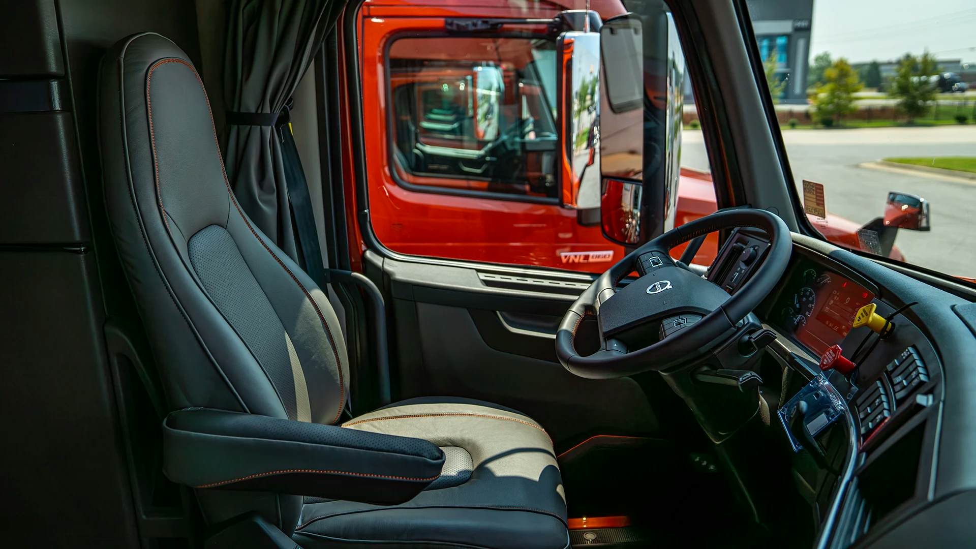 Interior of a modern and well-maintained semi-truck cabin, with a focus on the driver's seat, which has grey upholstery with orange stitching. The dashboard, featuring red-illuminated gauges and the Volvo logo on the steering wheel, is visible, indicating the vehicle's brand.
