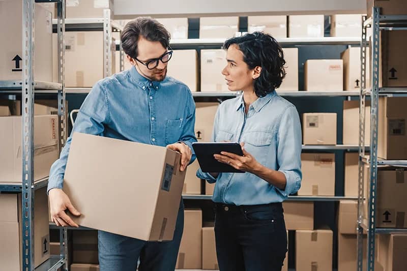 Two warehouse workers in casual blue attire are having a discussion in a storeroom. The man is holding a cardboard box while the woman, holding a digital tablet, appears to be giving instructions or sharing information. They are surrounded by shelves stocked with neatly organized boxes.