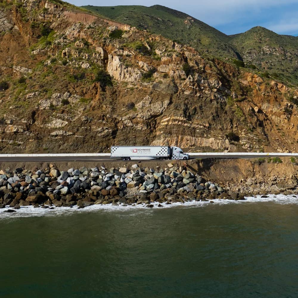 A white semi-truck from 'SCHWARZ LOGISTICS' is parked on the side of a coastal road, with the ocean on one side and steep, rugged cliffs on the other. The scene captures the beauty of transport routes that cut across diverse landscapes, under a clear blue sky.