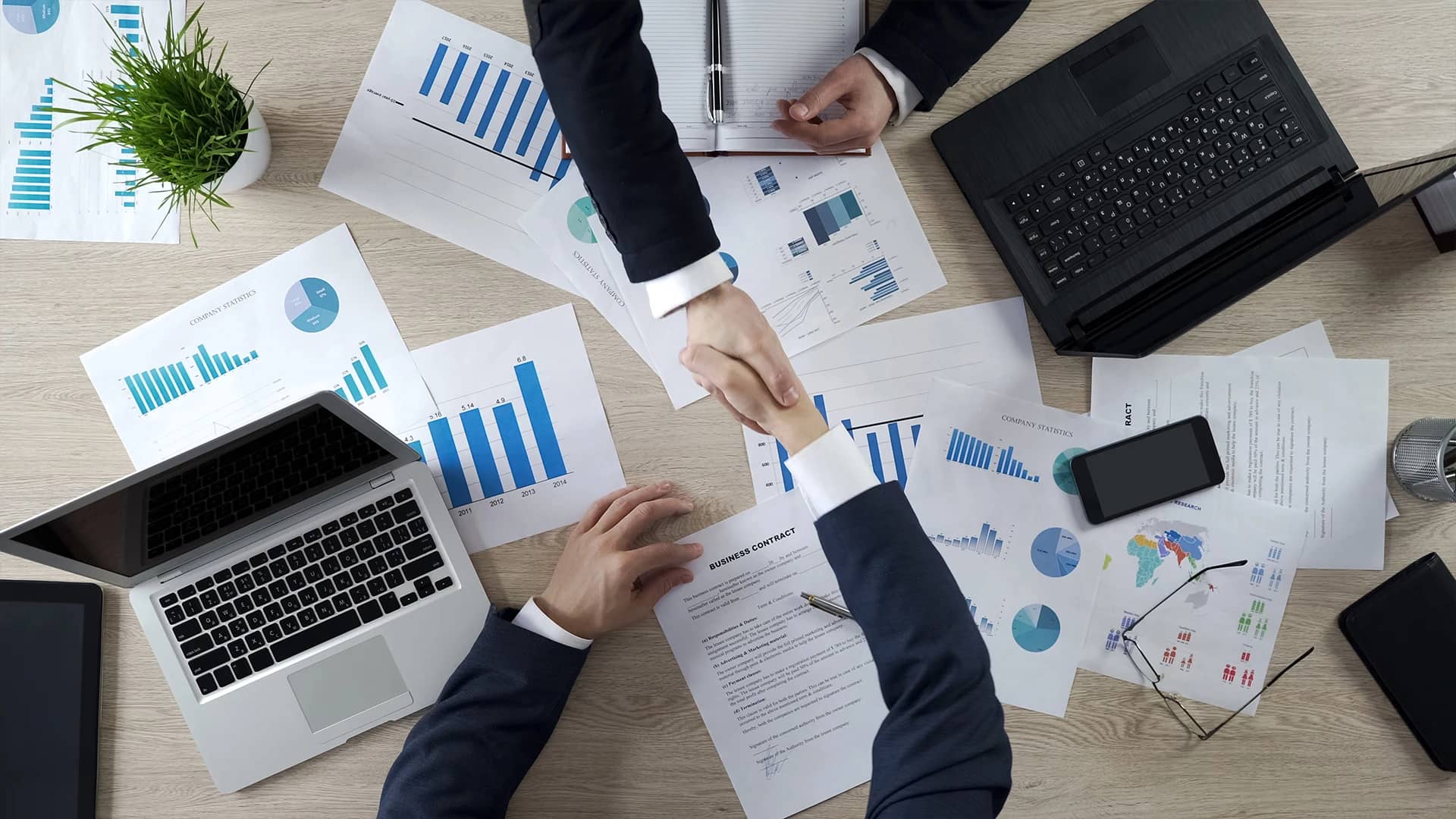 This image shows a top-down view of a business setting where two professionals are shaking hands over a desk, symbolizing a successful agreement or partnership. On the desk, there are various business documents, charts, and graphs scattered around, along with a laptop, a notebook, a pen, a smartphone, and a pair of glasses, indicating a thorough discussion of data or a strategic plan. The handshake is at the center, highlighting the importance of personal relationships in business dealings.