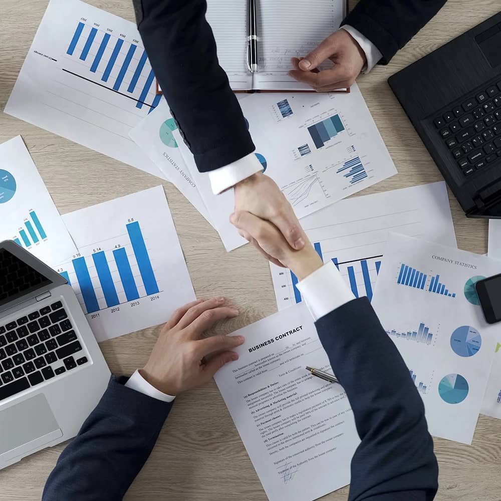 A top-down view of a professional setting where two individuals in business attire are shaking hands over a desk, indicating a successful negotiation or agreement. The desk is strewn with various documents, including a business contract, financial reports, graphs, and charts reflecting company statistics and performance metrics. A laptop, a notebook, and a smartphone are also on the desk, suggesting a contemporary, tech-enabled business environment. The handshake, centered in the composition.