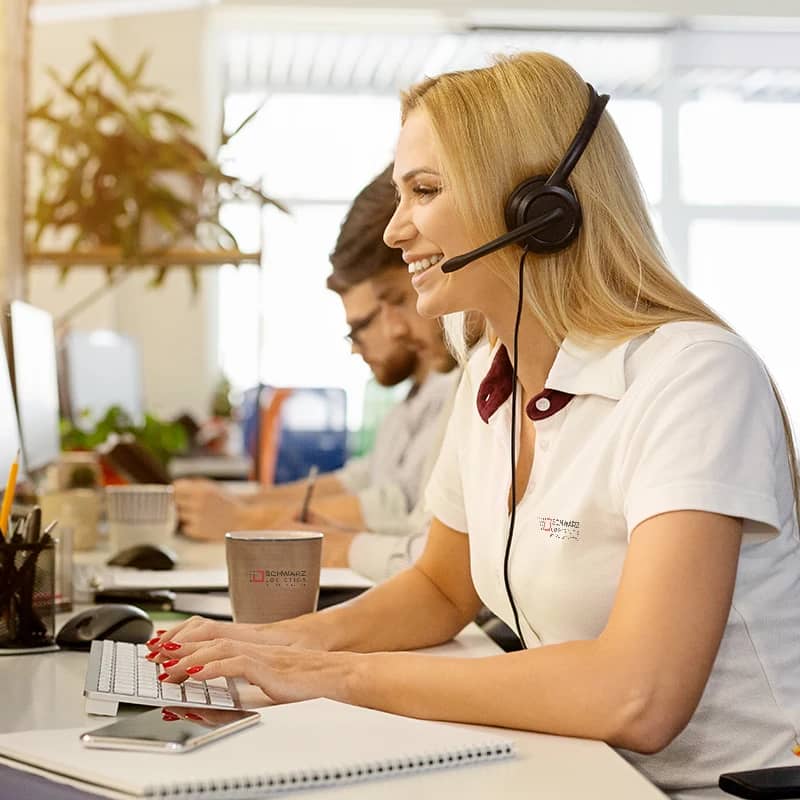 A cheerful customer service representative wearing a white polo shirt with the company logo "SCHWARZ LOGISTICS" on her left chest.