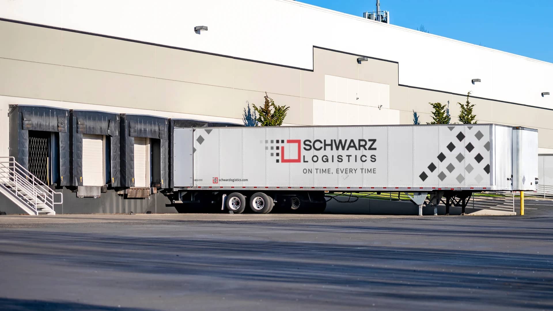 A large, white commercial building with multiple loading docks. Parked at one of the docks is a long white semi-trailer with the logo "SCHWARZ LOGISTICS" on its side, signifying that the facility is likely used for the distribution of goods. The trailer is backed into the dock, and loading dock doors are covered by black flexible curtains to seal the space between the dock and the trailer, indicating that loading or unloading activities may be taking place.