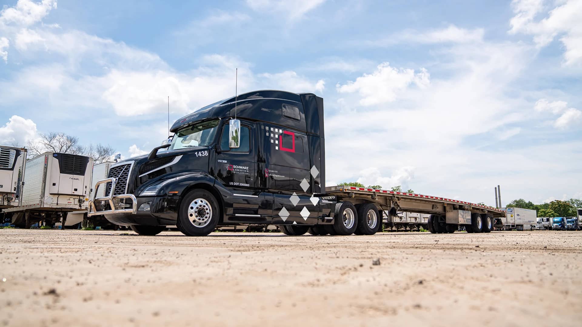 A semi-truck with a flatbed trailer, bearing the logo "Schwarz Logistics," parked in a sunny lot with other trucks.