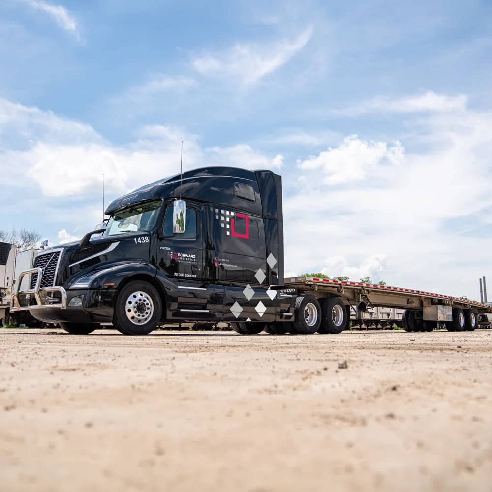 A sleek black flatbed semi-truck with the 'SCHWARZ LOGISTICS' logo on the side is parked on a concrete surface under a blue sky with scattered clouds. The truck appears robust and ready for hauling, with its flat trailer bed empty and visible, highlighting its cargo transport capabilities.