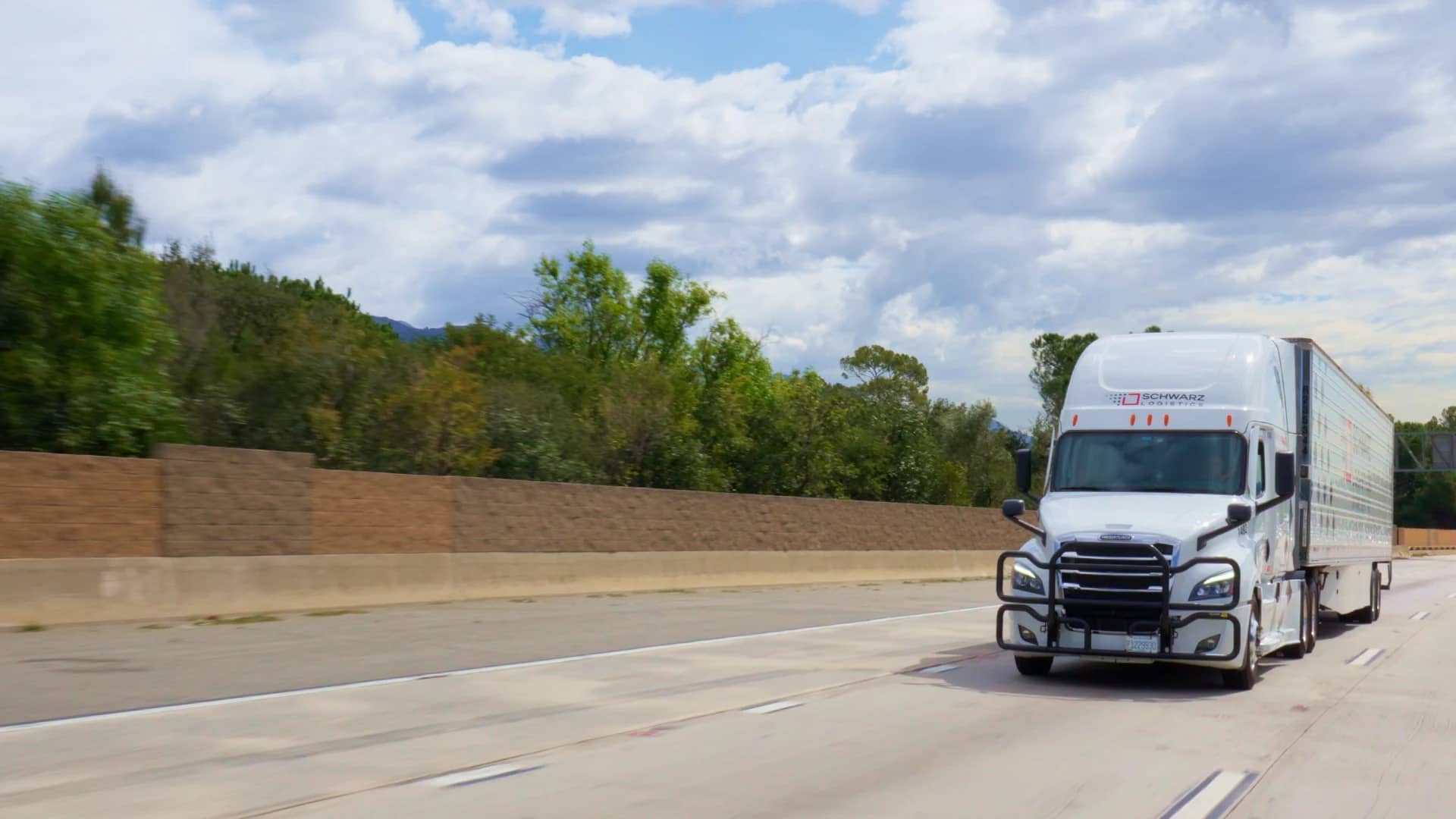 A white Freightliner Cascadia semi truck hauling a trailer travels down a straight, multi-lane highway.