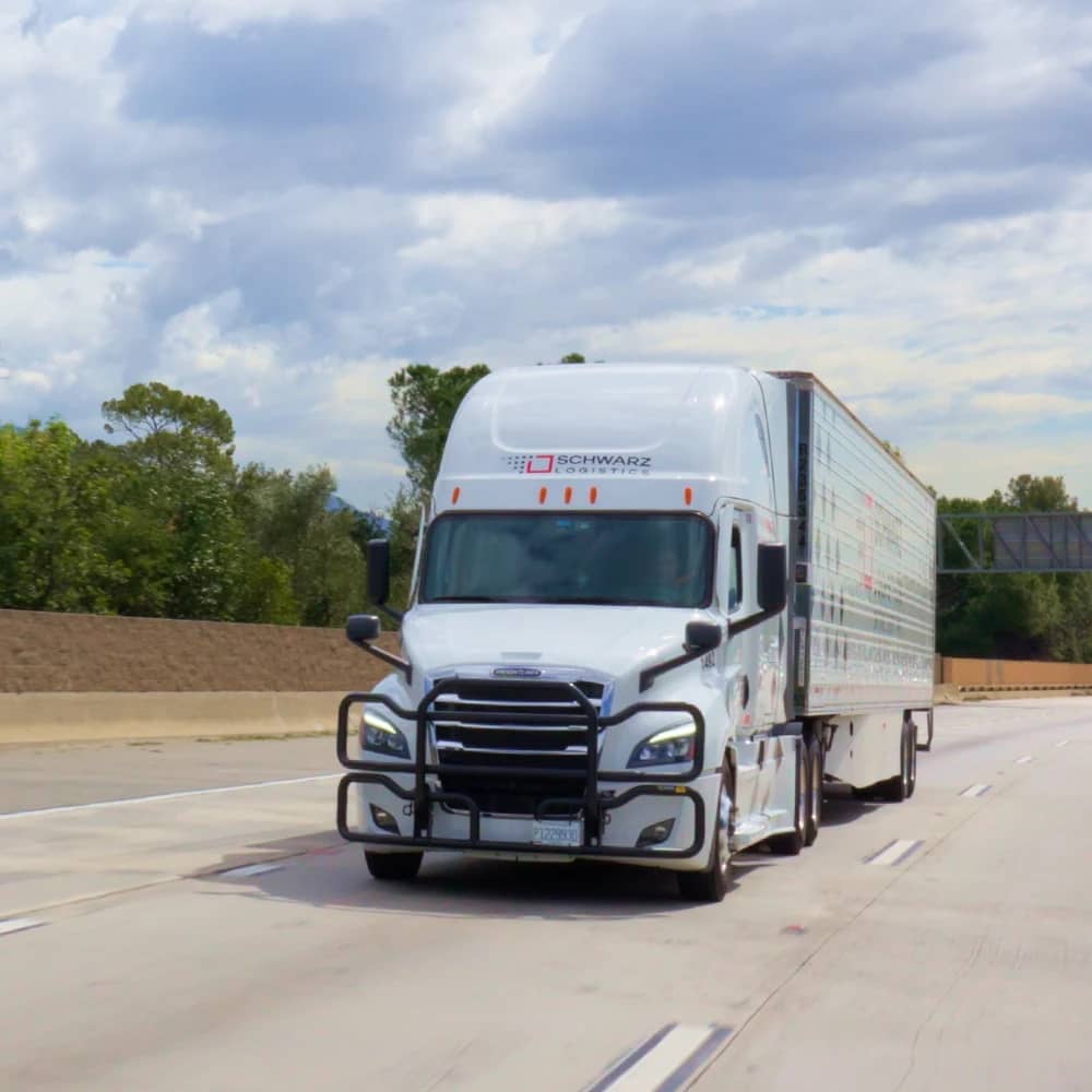 A white semi-truck with a large enclosed trailer, marked with the logo of "SCHWARZ LOGISTICS" on the side, is driving on a highway. The truck is in motion, as indicated by the slight blur of the scenery around it. The sky is partly cloudy, suggesting it might be a day with mixed weather.