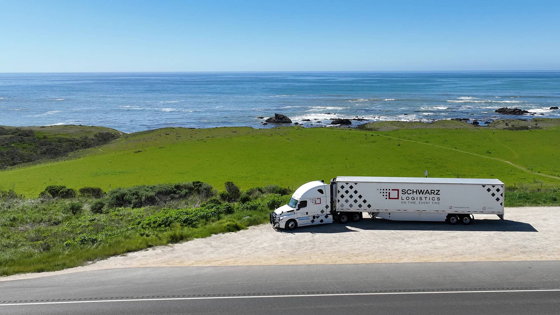 A semi-truck with a white tractor and a long trailer emblazoned with "SCHWARZ LOGISTICS" on the side, suggesting it is part of a logistics and transport company. The truck is parked on a roadside area with a lush green landscape and a scenic view of the sea.