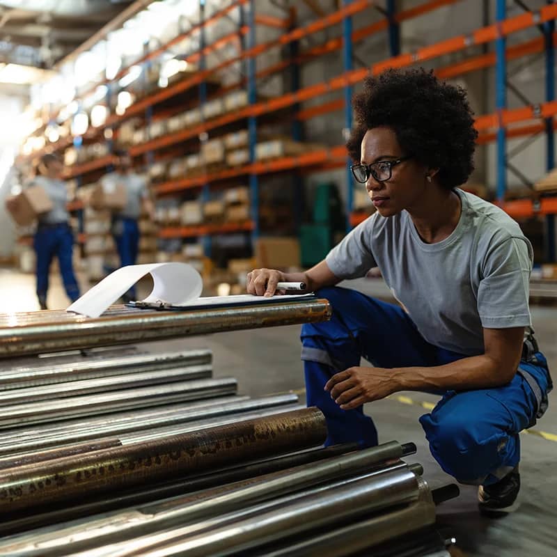 A focused worker in a warehouse inspects materials with a clipboard, with shelves stocked with boxes and colleagues working in the background.