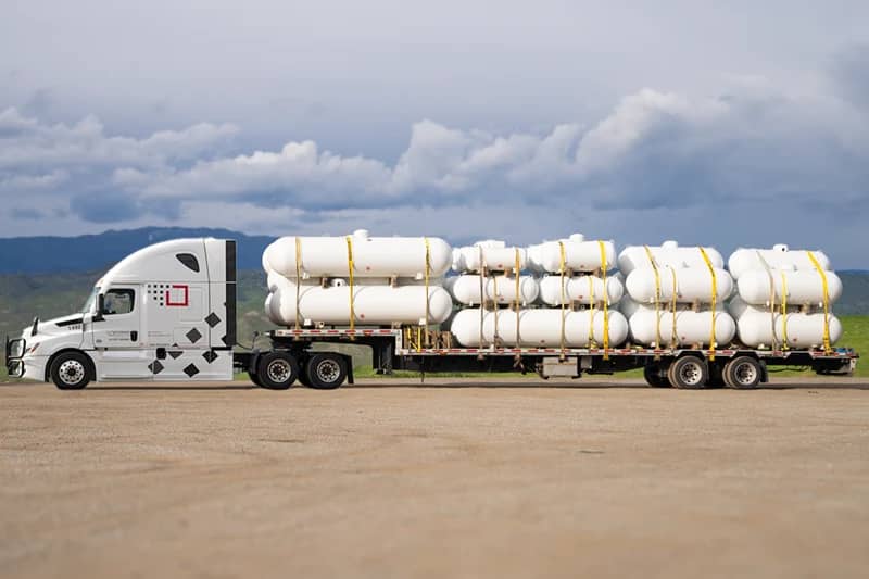 A modern white semi-truck connected to a step deck trailer, which is loaded with multiple large white tanks, each secured with yellow straps.