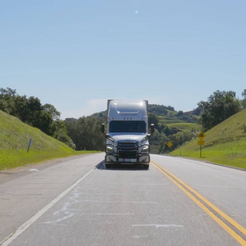 A large semi-truck driving on a two-lane road. The setting is idyllic with green hills on either side under a clear blue sky, suggesting a peaceful, rural area.