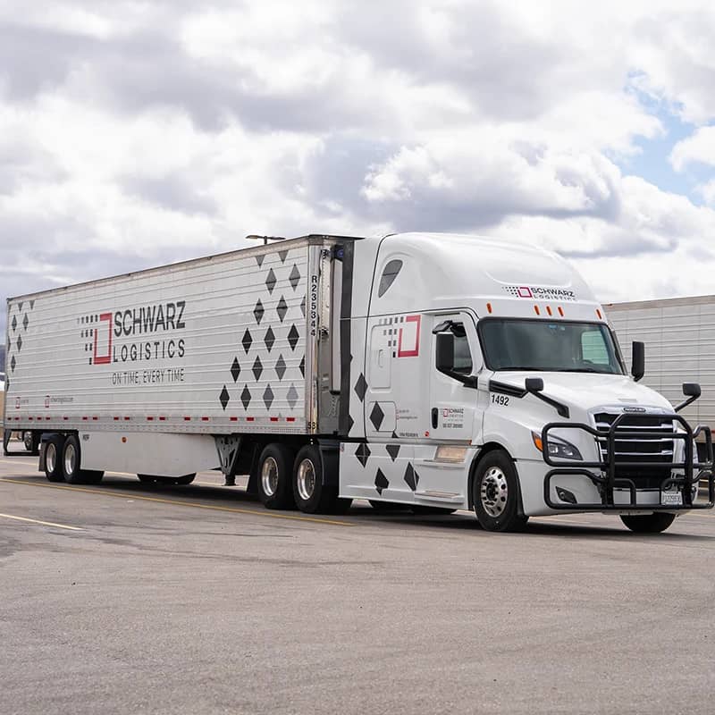 A "Schwarz Logistics" semi-trailer truck parked on a paved area, likely a loading dock or a parking lot, with industrial buildings in the background.