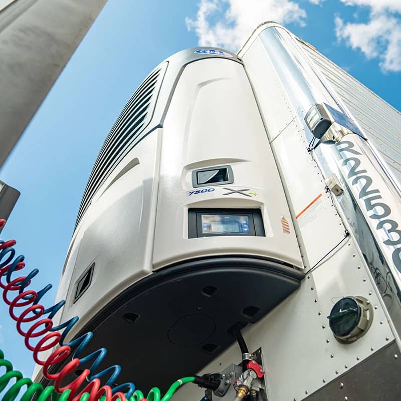 An upward view of a refrigeration unit on a trailer, set against a bright blue sky with scattered clouds. The unit, likely a part of a refrigerated transport vehicle, is branded with the Carrier logo and labeled with the model "7500 X", indicating it is a piece of specialized equipment used for controlling the temperature inside the trailer. The presence of coiled air lines and electrical cables suggests its integration with the vehicle's systems.