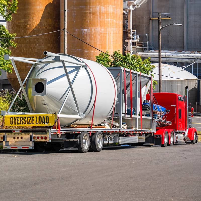 Red truck carrying oversized tank with "OVERSIZE LOAD" banner.