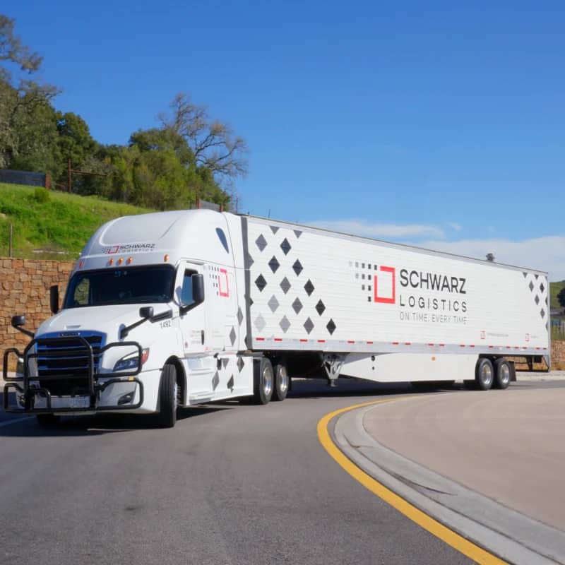 A white semi-truck with "SCHWARZ LOGISTICS" branding on the trailer is driving on the road, with a clear sky above and green trees in the background.