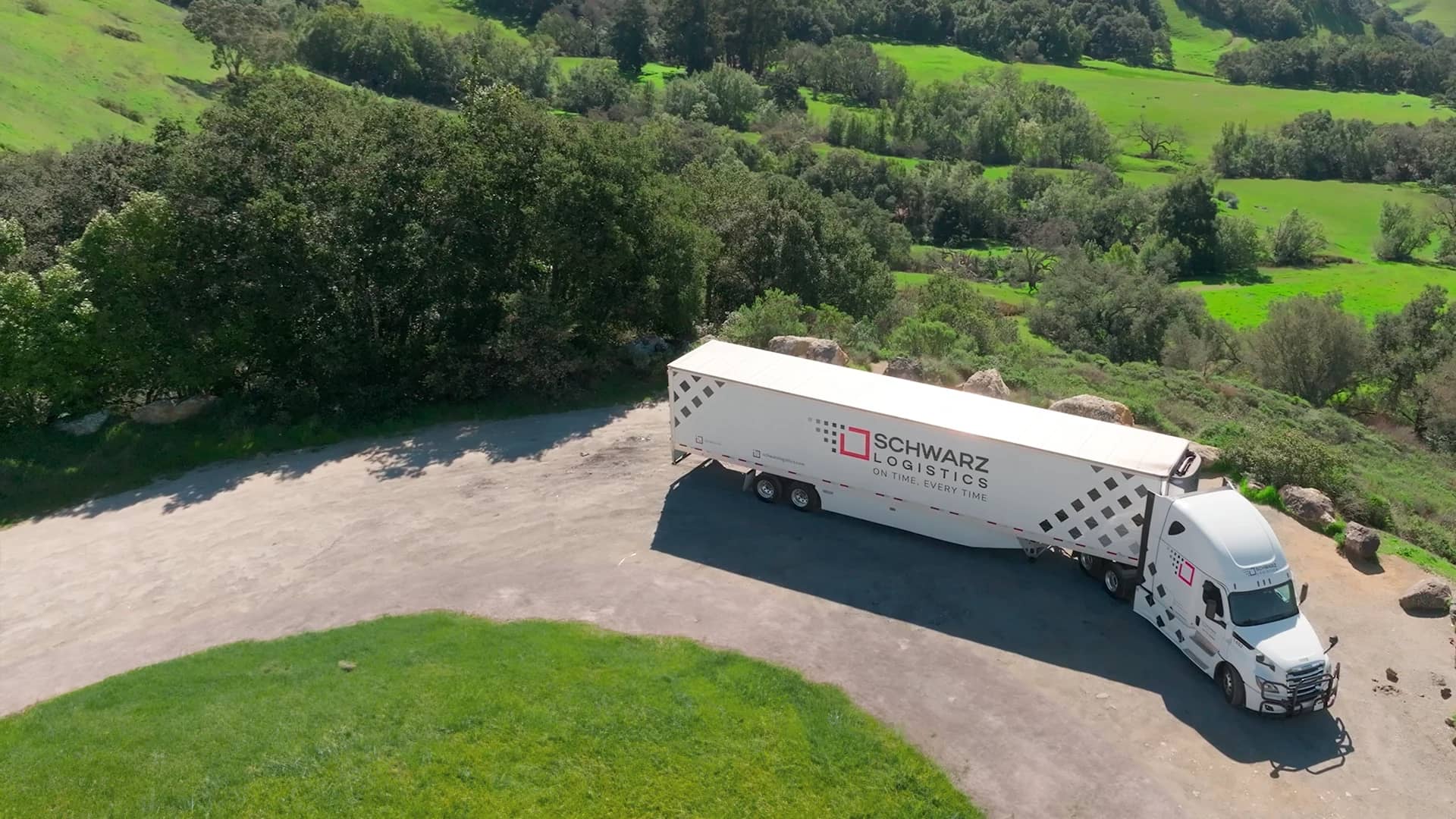 Aerial view of a white semi-truck with "SCHWARZ LOGISTICS" branding on the trailer parked on a gravel lot, surrounded by lush greenery and hills.