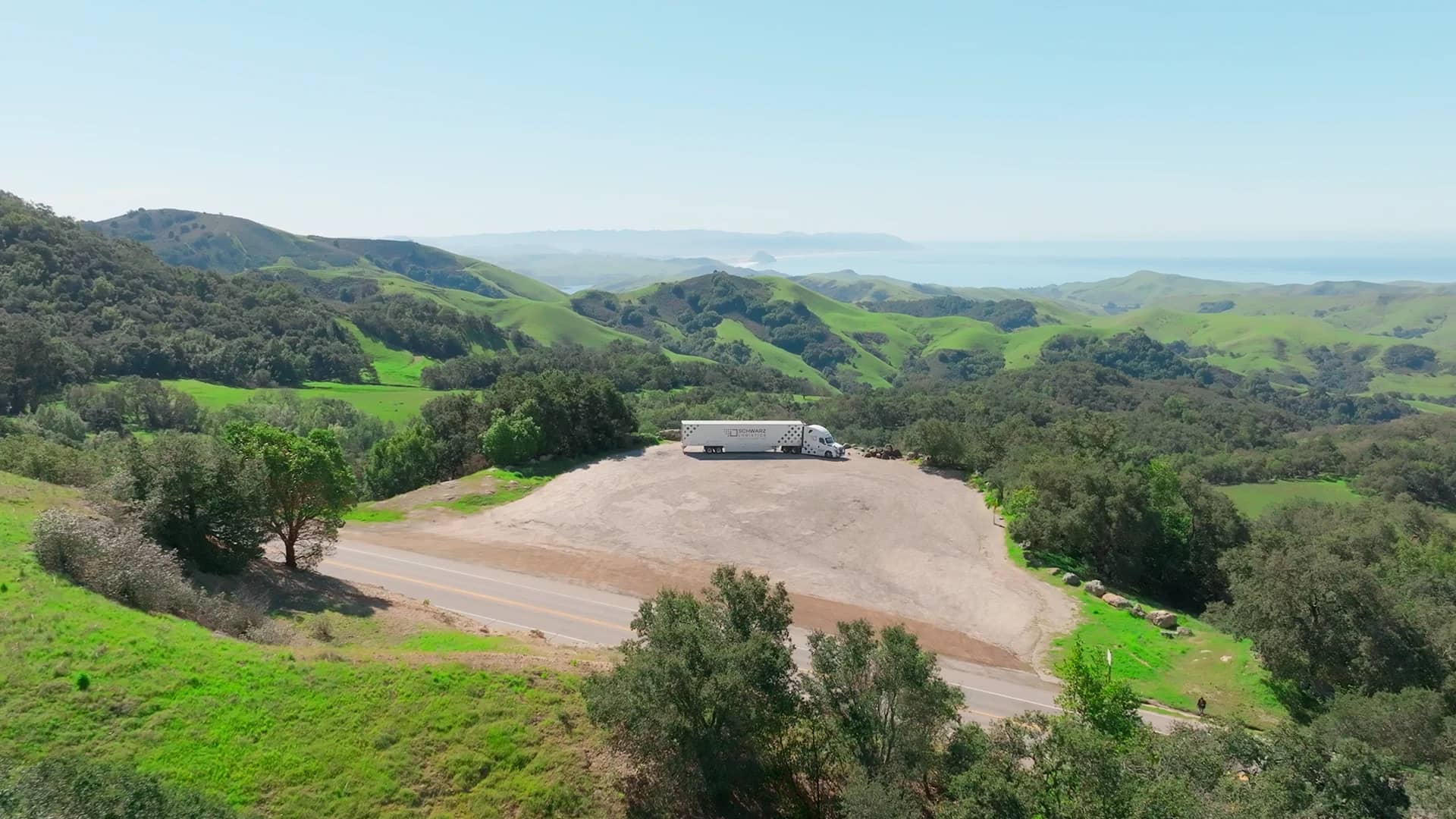 Pastoral landscape with lush green hills rolling into the distance under a clear blue sky. In the foreground, a large, white semi-truck with a patterned trailer is parked on a broad gravel turnout by a road that cuts through the scenery.