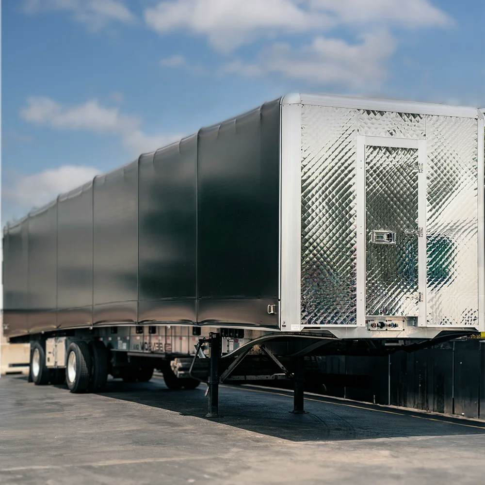 A close-up of the side and rear of a black Conestoga trailer with its distinctive rolling tarp system on a flatbed, showing the detailed texture of the tarp and the shiny diamond-patterned rear doors. The trailer is parked under a clear blue sky, indicating readiness for secure and versatile cargo transport.
