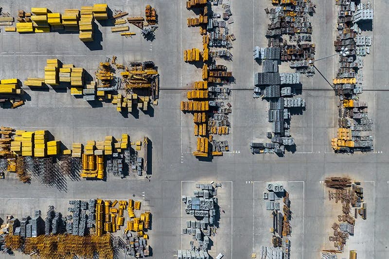 Aerial view of an industrial construction site showing neatly organized stacks of yellow and gray construction materials, including beams and pipes. The materials are methodically laid out across a large paved lot, divided by clear aisles, demonstrating order and planning in a large-scale operation.