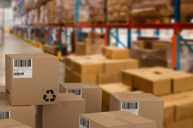 A close-up on a cluster of cardboard boxes with shipping barcodes and recycling symbols, focused in the foreground. The background features a defocused warehouse with tall shelving units stocked with various goods, portraying the dynamic environment of a distribution center for consumer packaged products.