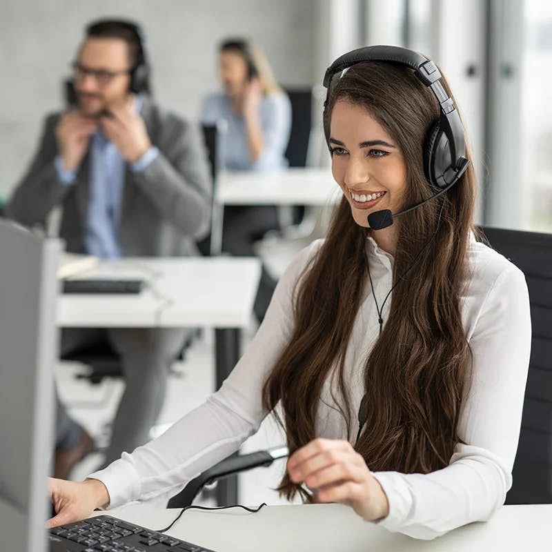 A cheerful customer support representative with a headset is engaged in conversation at her computer. Her focus is on the screen, suggesting active assistance to a client. In the softly blurred background, colleagues in a similar setup are also attending to calls, highlighting a busy and professional call center environment.