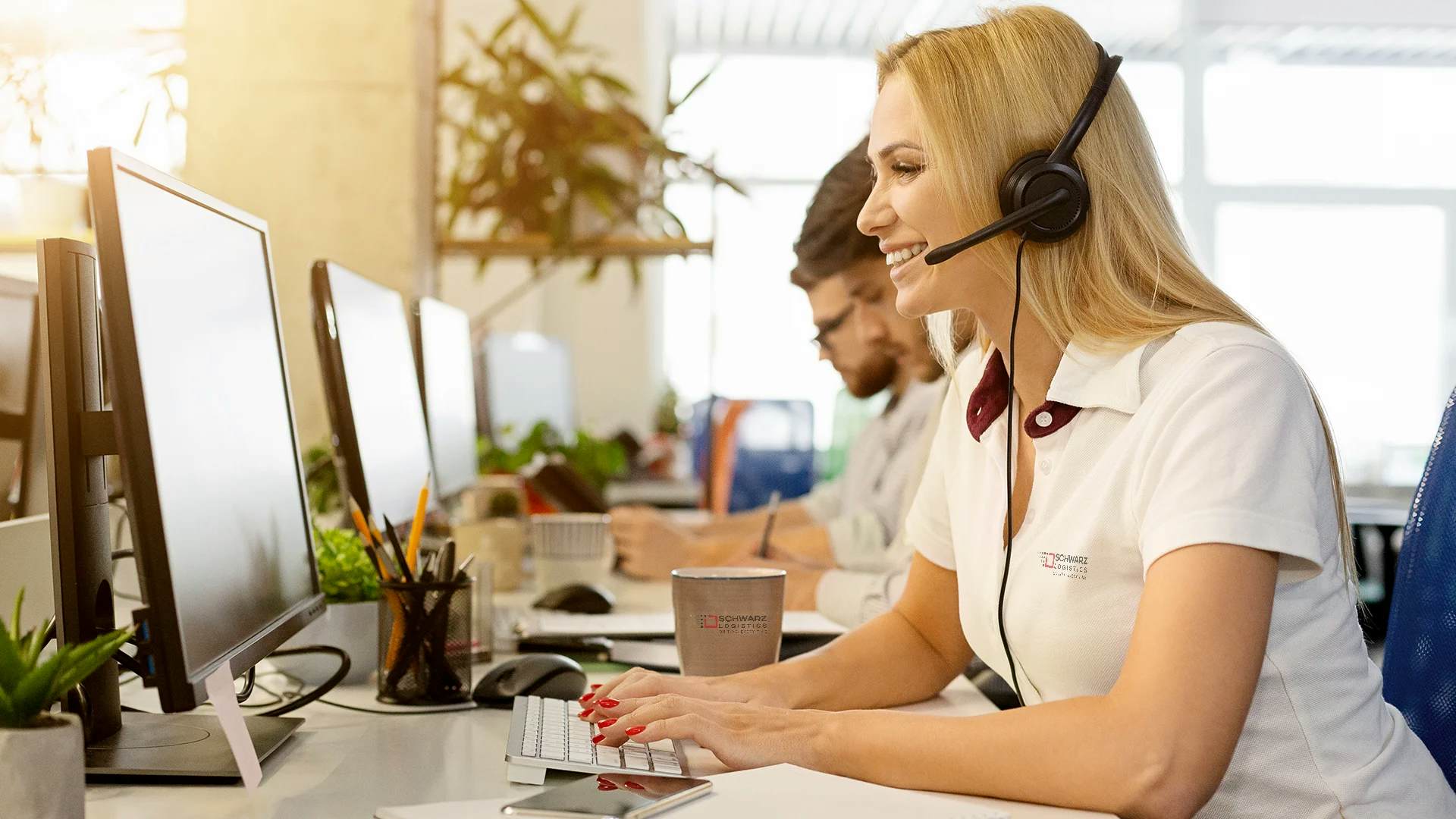 A smiling woman with a headset sits in an office environment, typing on a keyboard, indicative of a customer service representative. In the background, colleagues are also equipped with headsets and computers, focused on their work. The setting is bright with natural light, plants, and an atmosphere of professional engagement.
