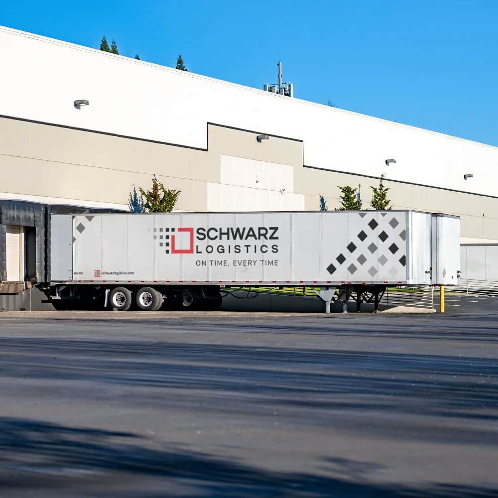 A large white semi-trailer truck with 'SCHWARZ LOGISTICS' branding is parked at a warehouse loading dock. The trailer features a geometric diamond pattern and the slogan 'ON TIME, EVERY TIME,' suggesting a focus on reliable and punctual delivery services.