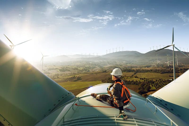 A technician in a safety harness sits atop a large wind turbine, overlooking a vast wind farm bathed in sunlight. The scenic landscape includes rolling hills and additional turbines stretching into the distance, symbolizing renewable energy and sustainable technology.