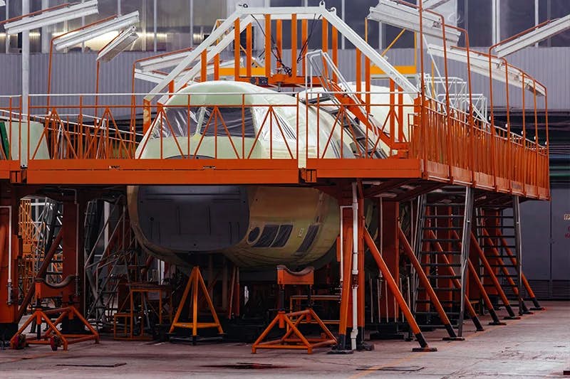 A partially assembled airplane within an industrial hangar, supported by various metal stands and scaffolding. The front section of the aircraft is visible, featuring a green primer coating, with an orange access structure surrounding it to allow engineers to work at different heights around the fuselage.
