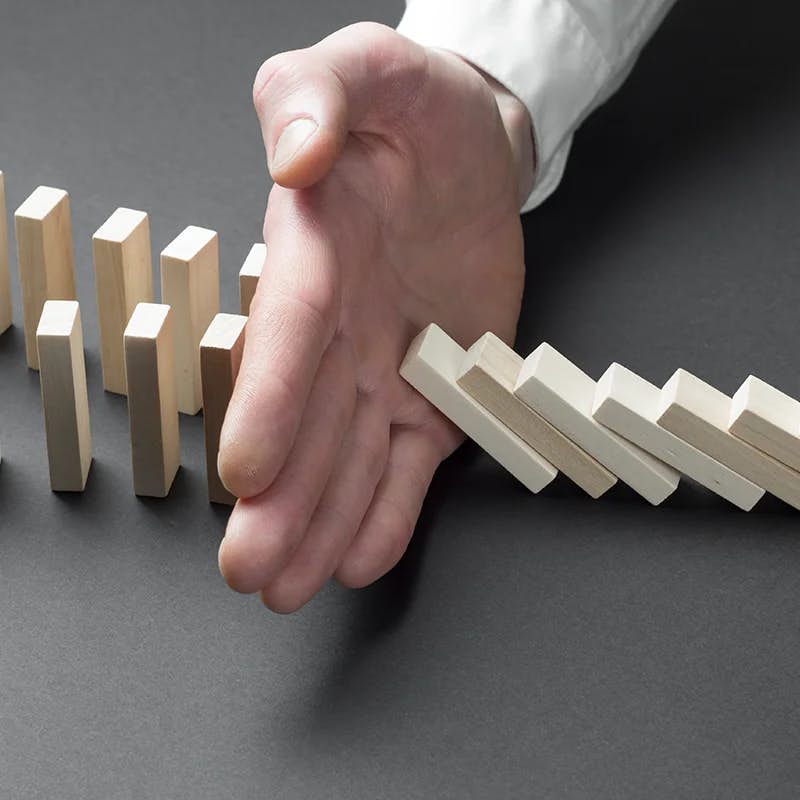 A person's hand is poised to stop a series of wooden dominoes from falling, with the dominoes arranged in a descending line on a gray surface. The hand, belonging to an individual wearing a white sleeve, symbolizes intervention or prevention of a potential chain reaction of events.