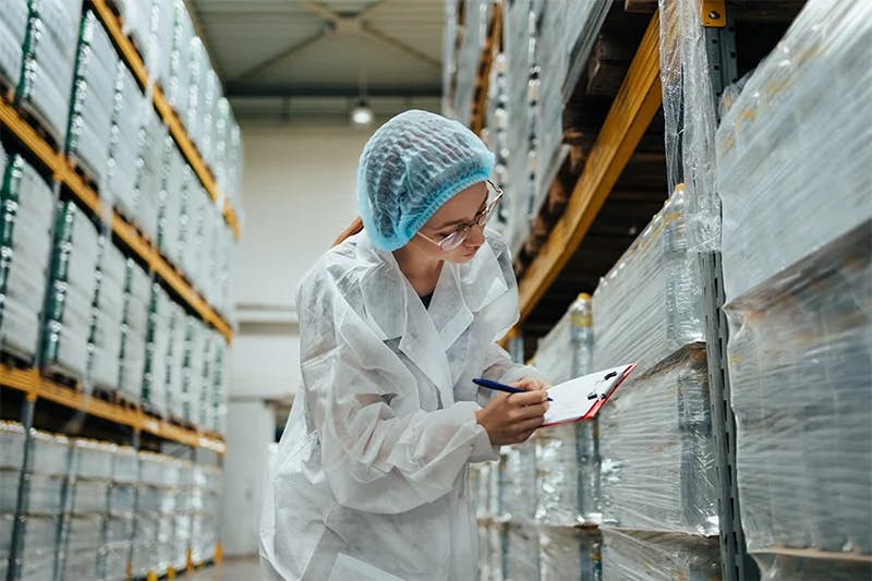 A focused worker in a white lab coat and blue hairnet is inspecting inventory in a pharmaceutical warehouse. She's writing on a clipboard while examining products on shelves, which are wrapped in protective plastic, emphasizing a controlled and hygienic environment.