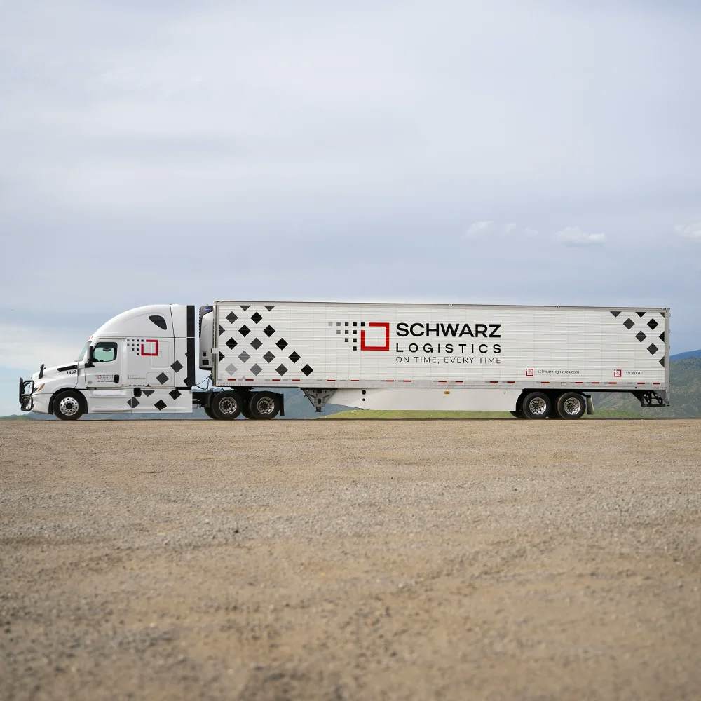 A white semi-truck with a large refrigerated trailer parked on a gravel lot, featuring the 'SCHWARZ LOGISTICS' branding along its side. The truck's trailer is adorned with a diamond pattern and the slogan 'ON TIME, EVERY TIME,' set against a backdrop of a cloudy sky and distant hills.