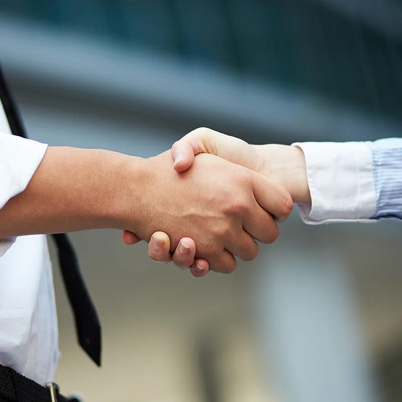 Close-up of a firm handshake between two individuals against a blurred background. One person is wearing a white shirt and the other a black sleeve, symbolizing a professional agreement or partnership.