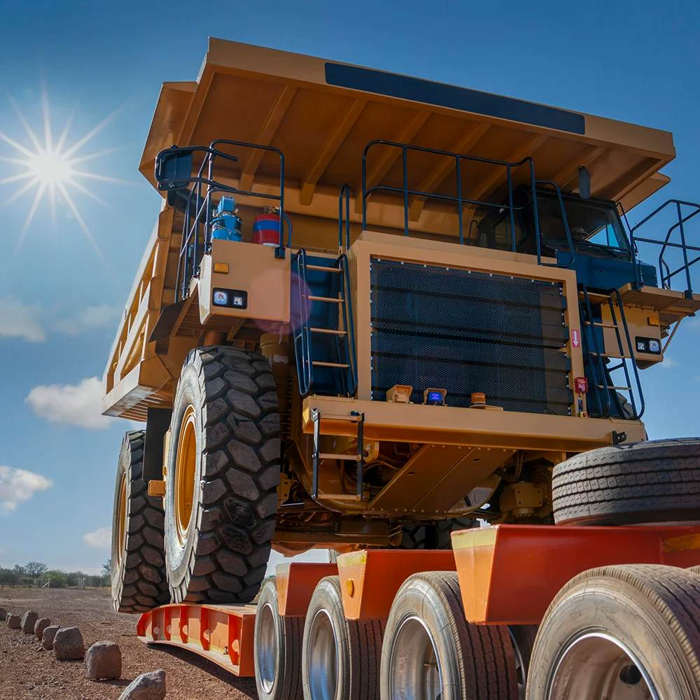A towering, massive yellow mining dump truck being transported on a red and gray multi-axle lowboy trailer under a sunny sky. The sheer scale of the truck is highlighted by the large tires that are almost the height of the trailer itself, emphasizing the specialized nature of heavy equipment transportation.