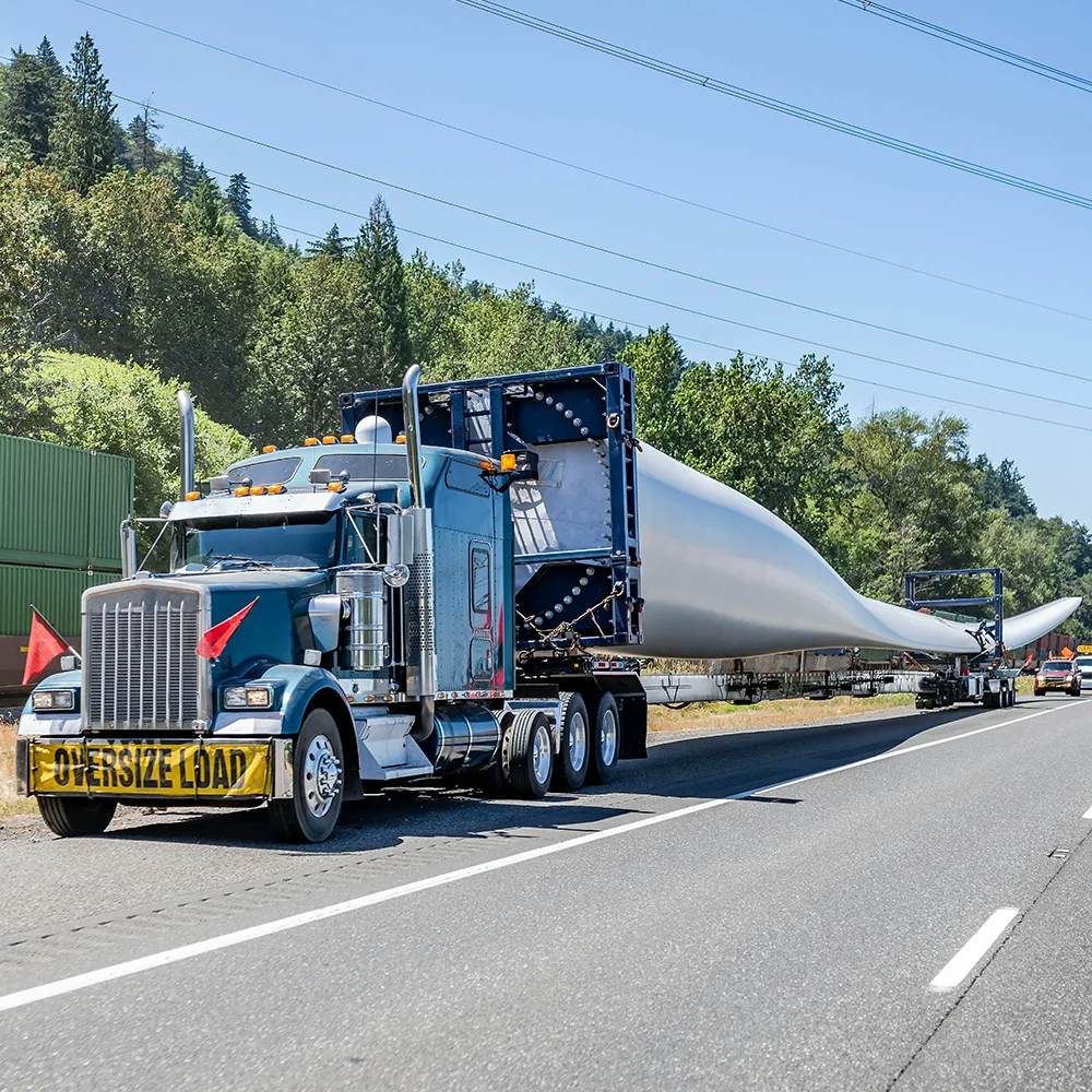 A blue classic semi-truck with chrome detailing is hauling an extremely long and oversized wind turbine blade on an extended flatbed trailer, marked by 'OVERSIZE LOAD' signage. The truck is on a highway with forested hills in the background, under a clear blue sky, indicating the transportation of renewable energy components.