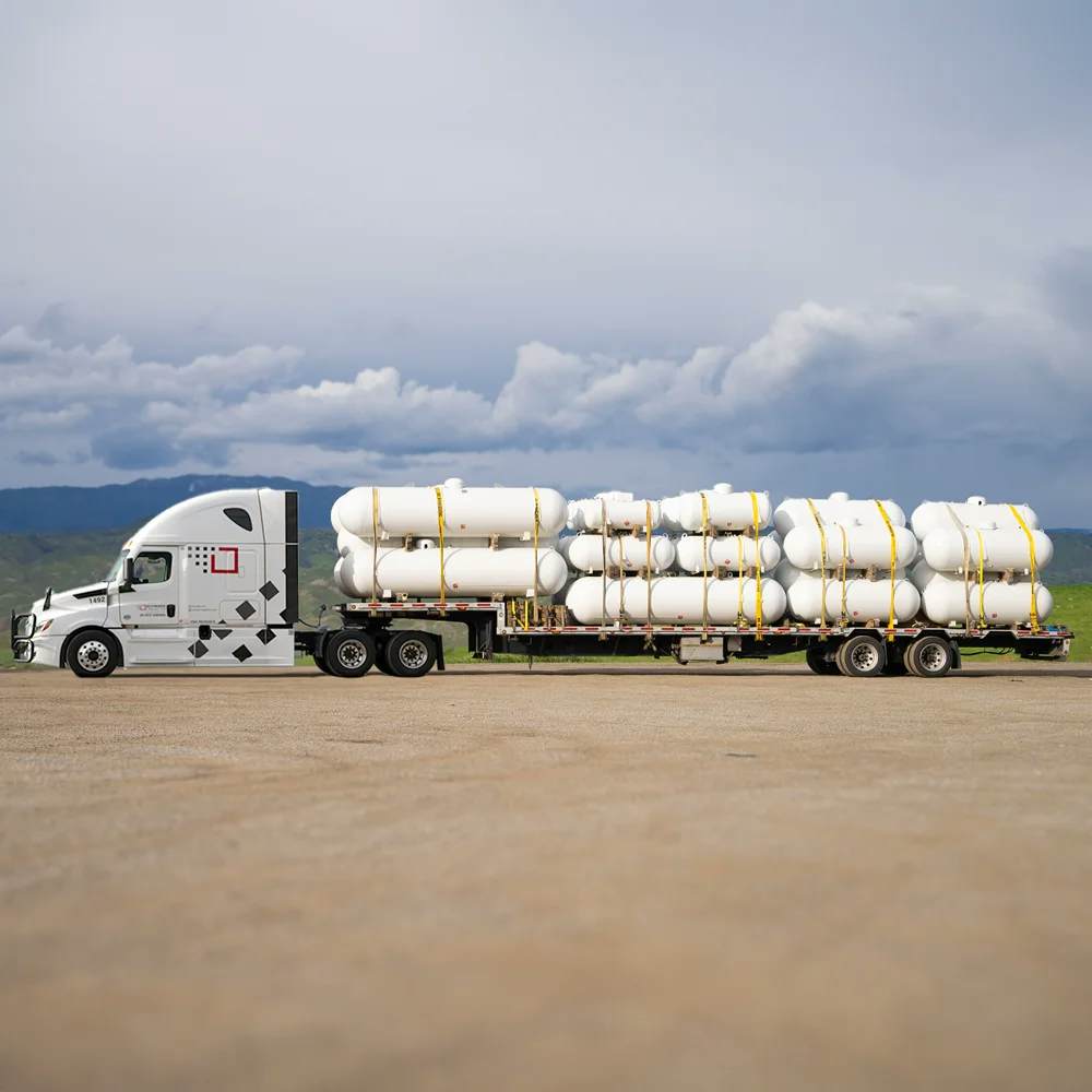 A white semi-truck with a step-deck trailer loaded with multiple large white cylindrical storage tanks secured with yellow straps, positioned against a backdrop of a dramatic sky and rolling hills, indicating readiness for specialized transport.