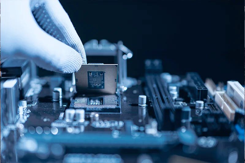 A gloved hand holds a microprocessor chip above a motherboard, highlighting the intricate details of semiconductor manufacturing. The focus is on the central processing unit with its pins ready to be inserted into the socket, set against the blurred backdrop of other electronic components on the board, all bathed in a cool blue light.