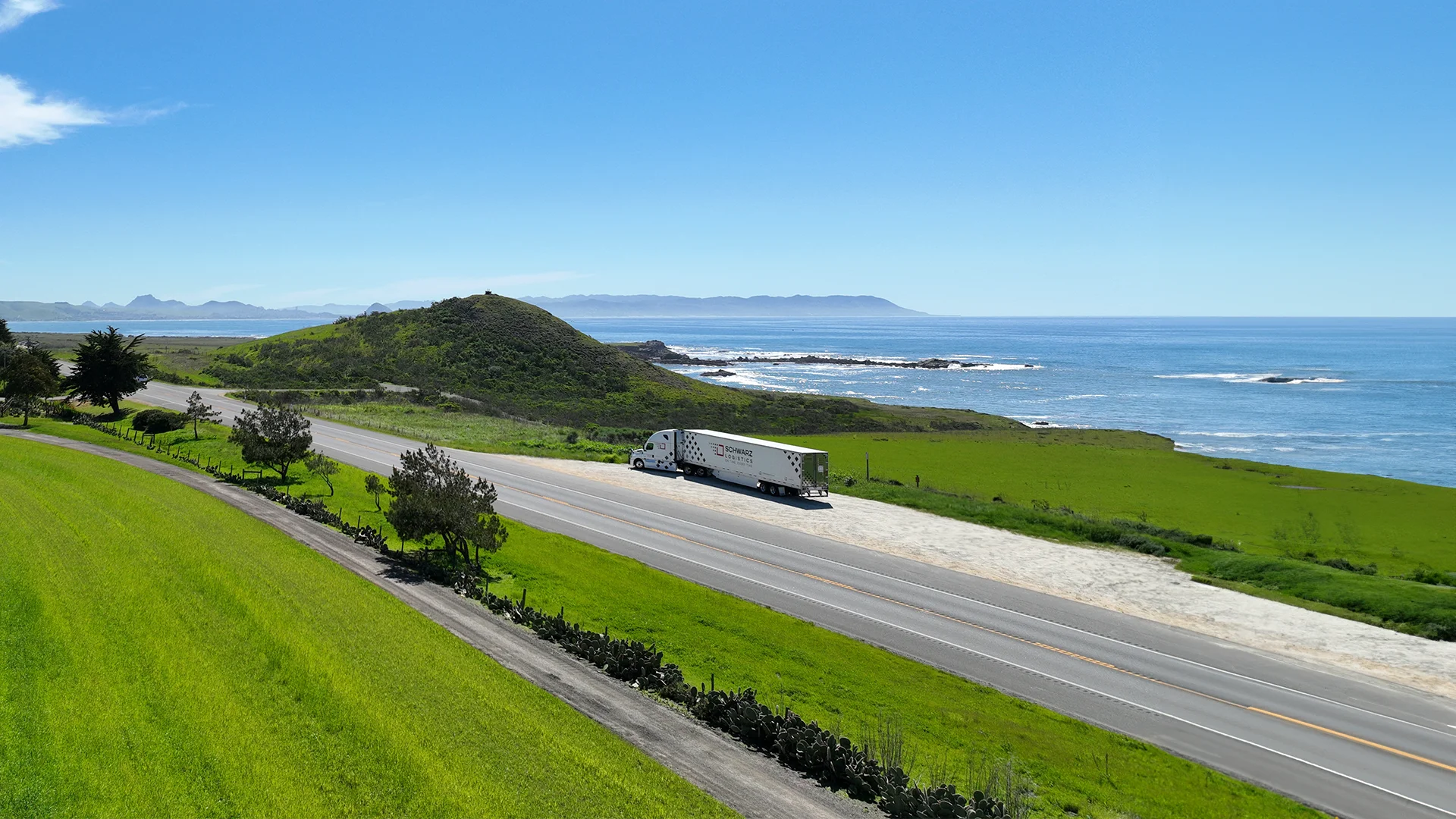 A panoramic view of a coastal highway with a white Schwarz Logistics commercial truck parked along the road. The scene includes lush green hills on the left and a clear blue ocean with rocky shores on the right, under a bright blue sky. In the distance, mountain silhouettes can be seen against the horizon, contributing to the serene and picturesque landscape.