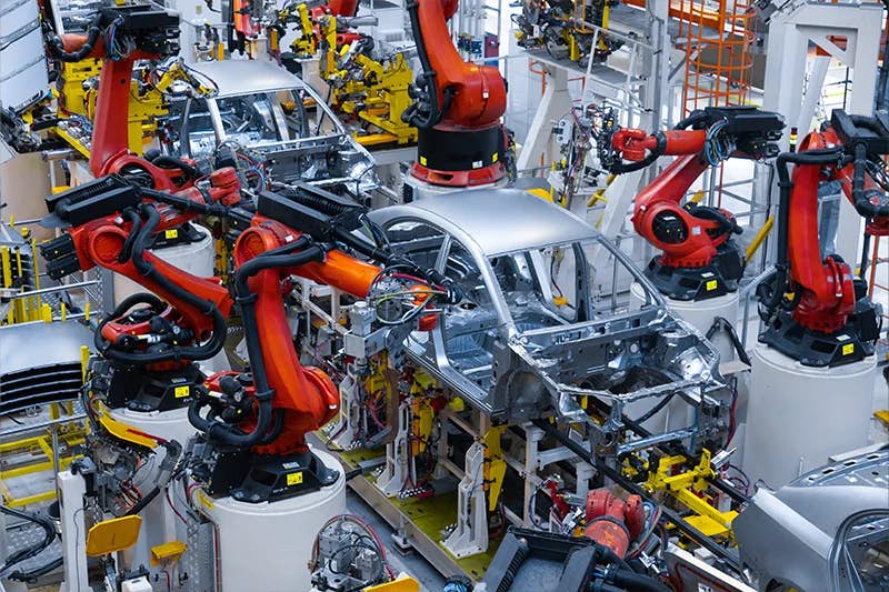 An automotive manufacturing line with multiple vibrant orange robotic arms working on assembling silver car bodies. The robots are equipped with various tools, situated in a highly organized and precise arrangement, demonstrating advanced automation in vehicle production.