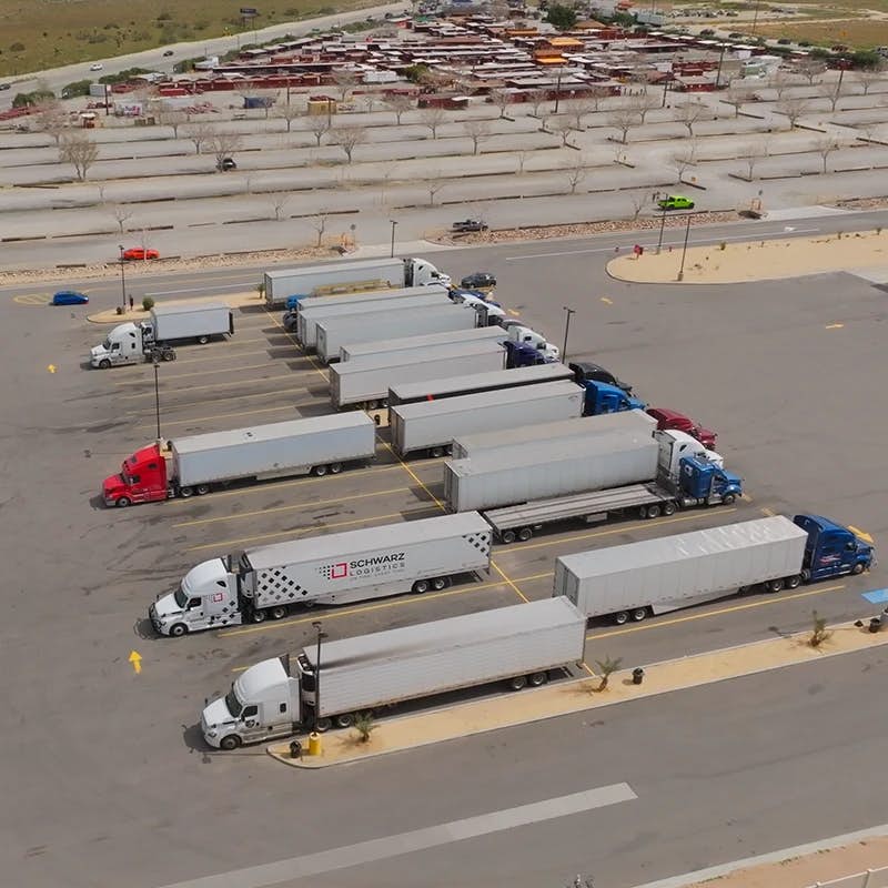 Aerial view of a logistics and transportation facility, showcasing a fleet of semi-trailer trucks parked in designated spots in a large, organized parking lot. Some trucks are connected to trailers, indicating preparedness for deliveries or pickups, while others are detached, possibly waiting for their next assignment. The variety of truck colors adds diversity to the scene. The facility appears to be in a spacious, possibly arid location given the presence of sand and sparse vegetation.