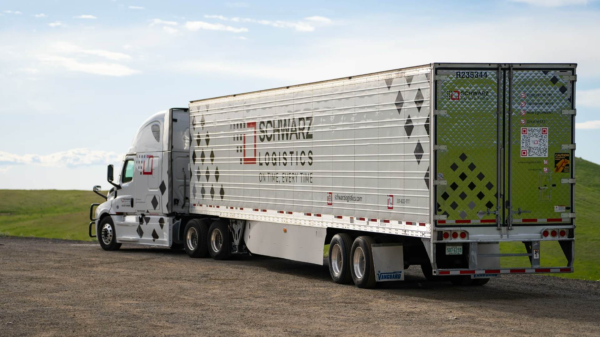 A semi-trailer truck parked on a gravel lot with a countryside landscape in the background.