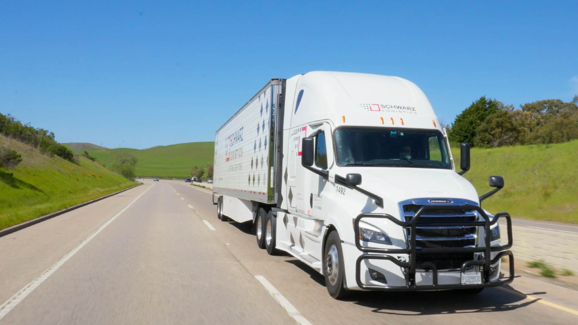 A white Freightliner Cascadia semi truck hauling a trailer travels down a straight, multi-lane highway.
