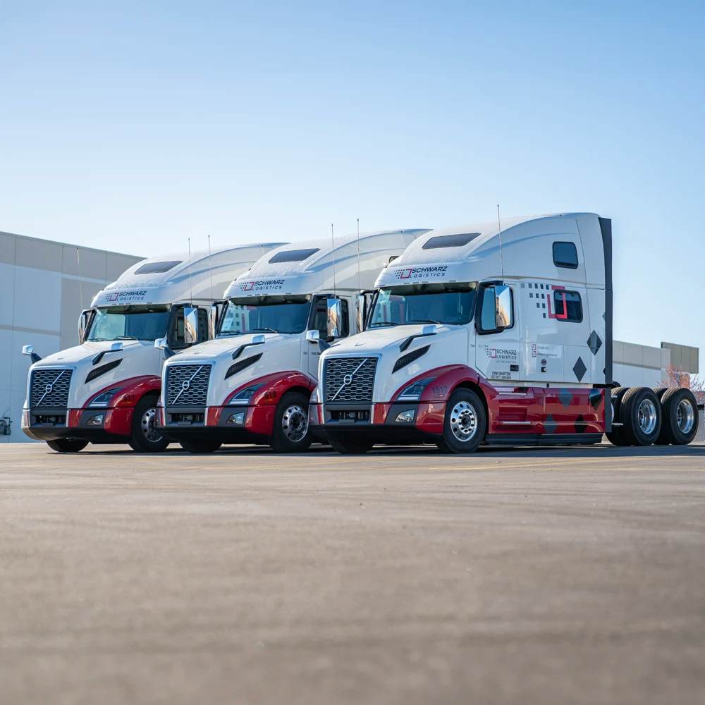 A fleet of three identical modern semi-trucks parked in a formation on a clear day. Each truck has a distinctive white cab with a striking red and white design on the front, accentuating the aerodynamic shape of the vehicles. The trucks bear the "SCHWARZ LOGISTICS" branding along with a checkered motif and a red stripe running along the bottom of the cabs. They are parked in front of a large industrial building, suggesting they are at a loading dock or distribution center.