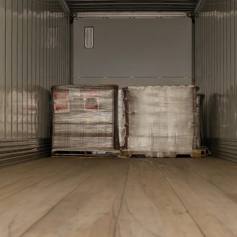 Interior of a dry van trailer with wooden flooring. There are two industrial pallets wrapped in plastic shrink wrap; the one on the left appears slightly disheveled with its contents partially visible, while the one on the right is more intact. The interior walls of the trailer have horizontal grooves typical of freight trailers, which are often used to secure goods during transport. The scene suggests that the trailer is either in the process of being loaded or unloaded.