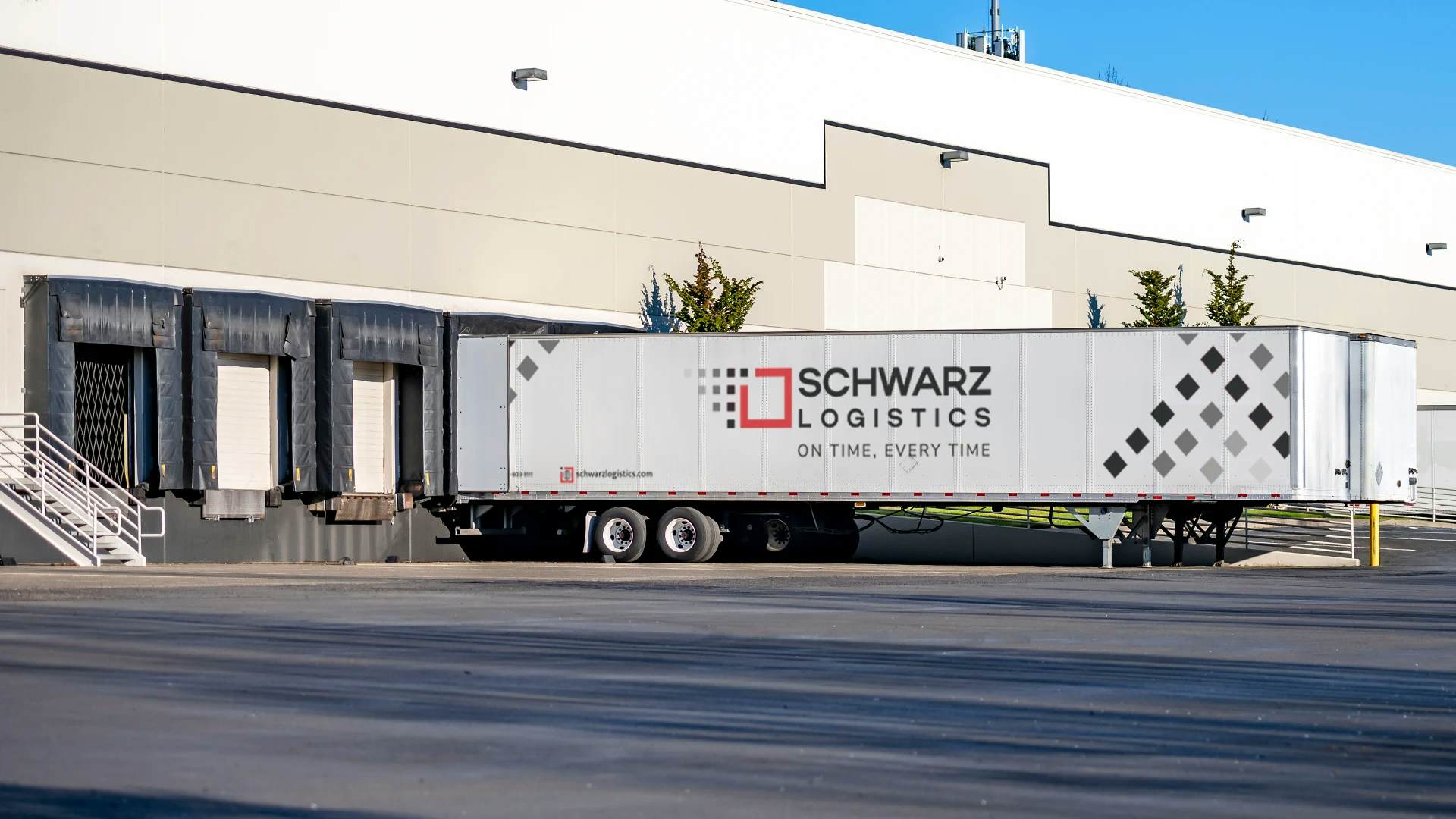 A large, white commercial building with multiple loading docks. Parked at one of the docks is a long white semi-trailer with the logo "SCHWARZ LOGISTICS" on its side, signifying that the facility is likely used for the distribution of goods. The trailer is backed into the dock, and loading dock doors are covered by black flexible curtains to seal the space between the dock and the trailer, indicating that loading or unloading activities may be taking place.