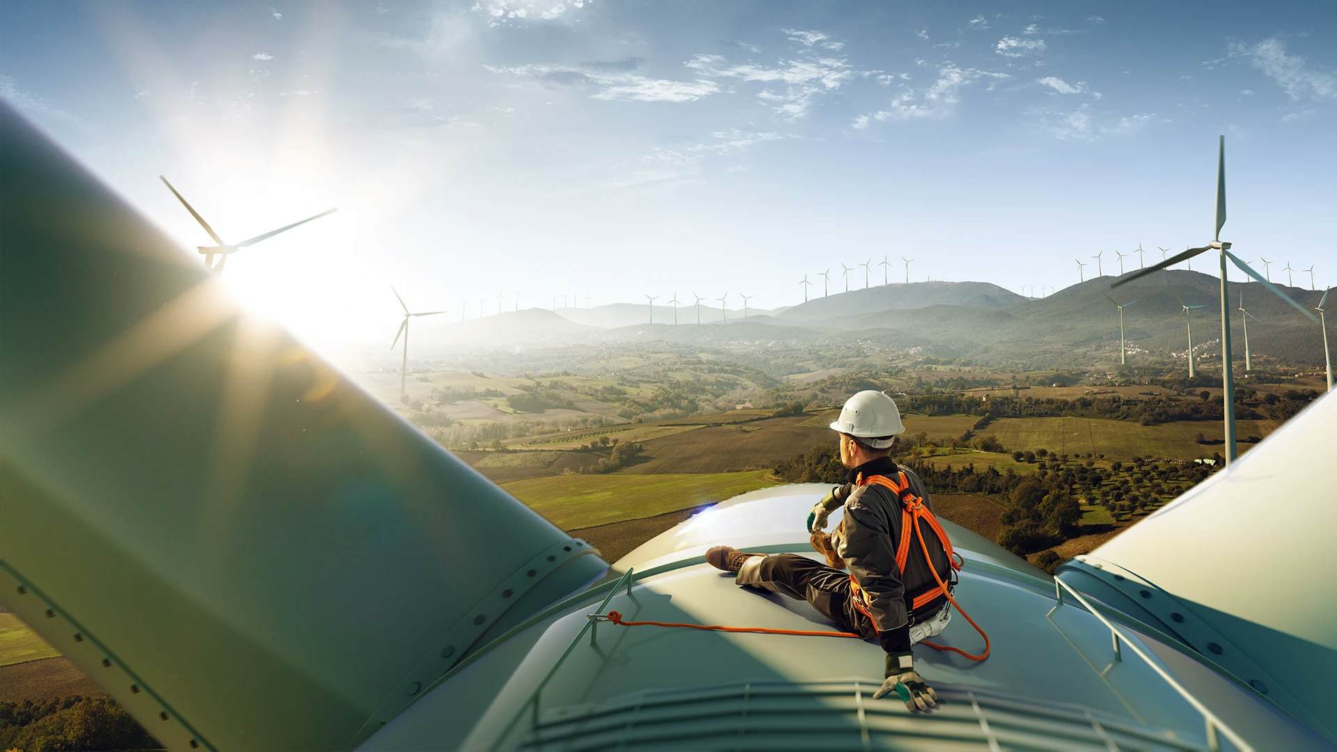 Technician sitting on a wind turbine with a scenic view of wind farm and hills.