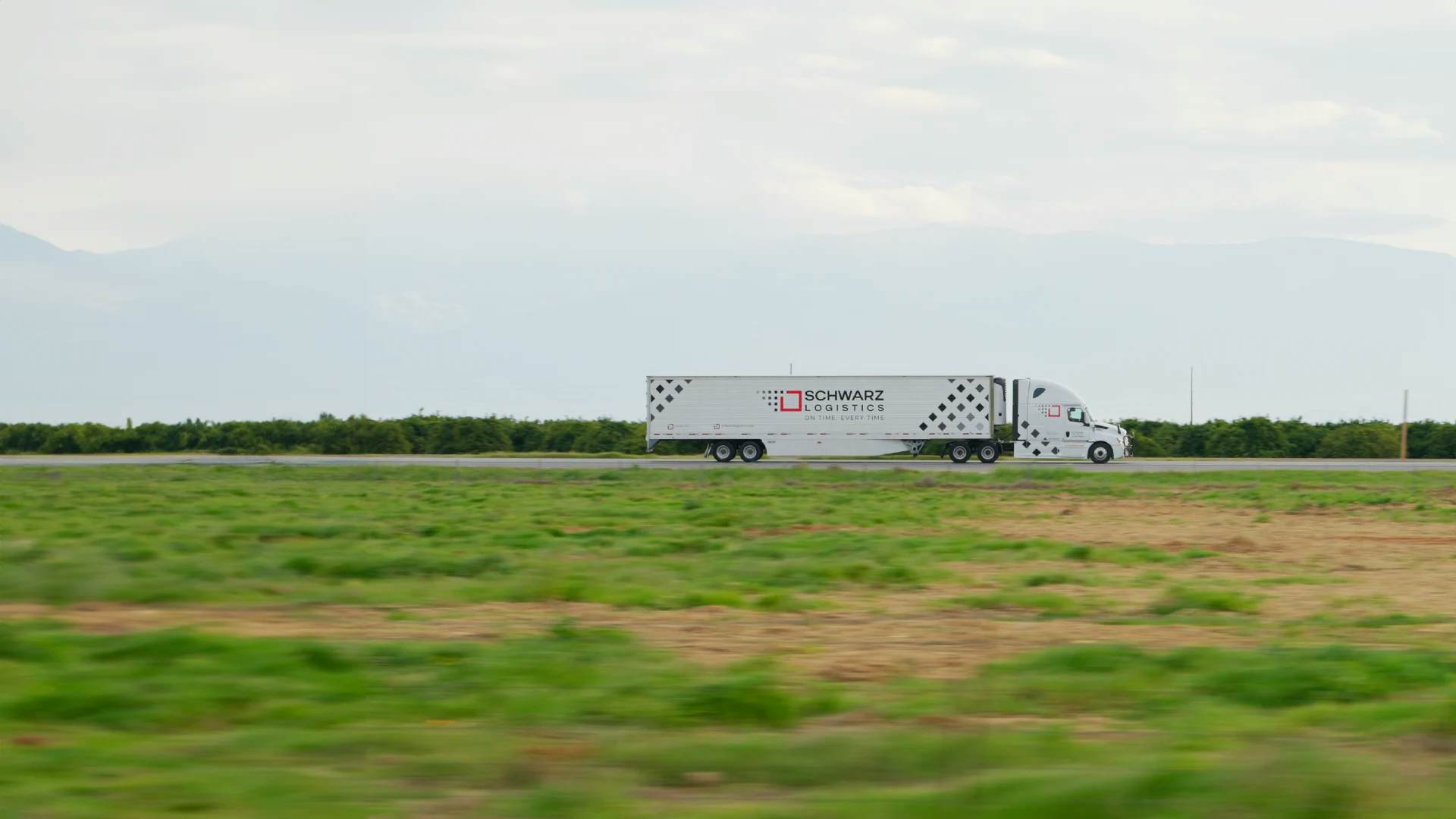 A 'SCHWARZ LOGISTICS' semi-truck is in motion on a rural road, with a clear focus on the moving vehicle against a blurred background of green fields and distant mountains, suggesting speed and efficiency in transportation services.