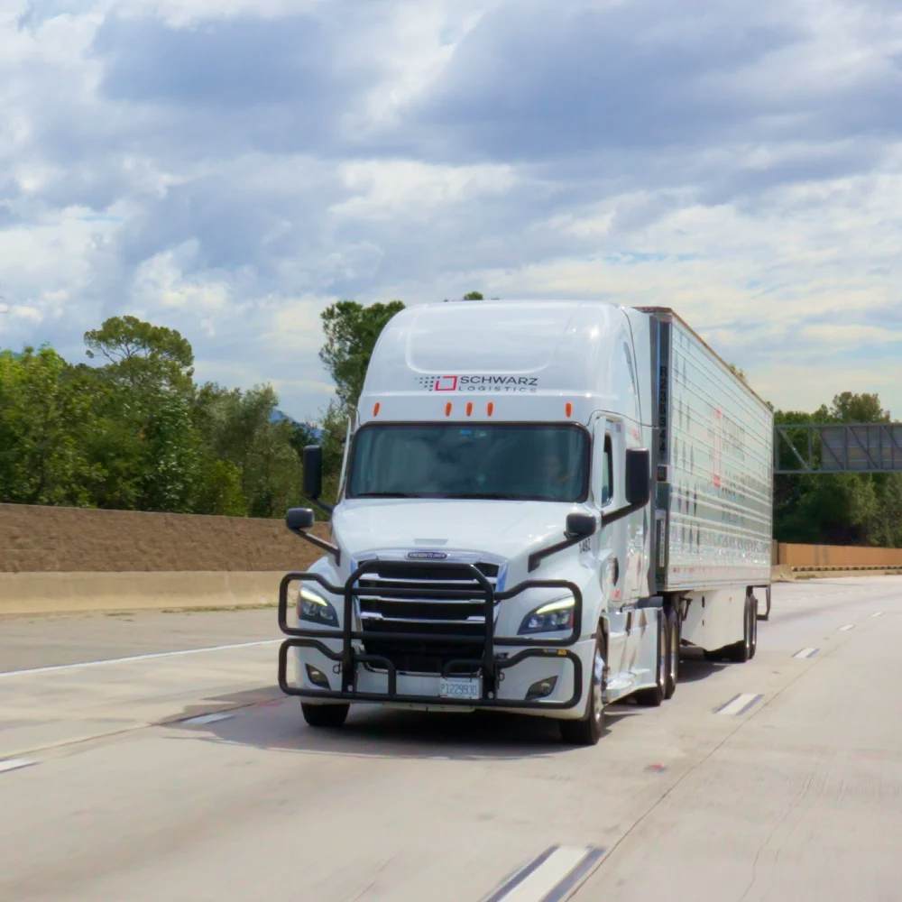 A white semi-truck with a large enclosed trailer, marked with the logo of "SCHWARZ LOGISTICS" on the side, is driving on a highway. The truck is in motion, as indicated by the slight blur of the scenery around it. The sky is partly cloudy, suggesting it might be a day with mixed weather.