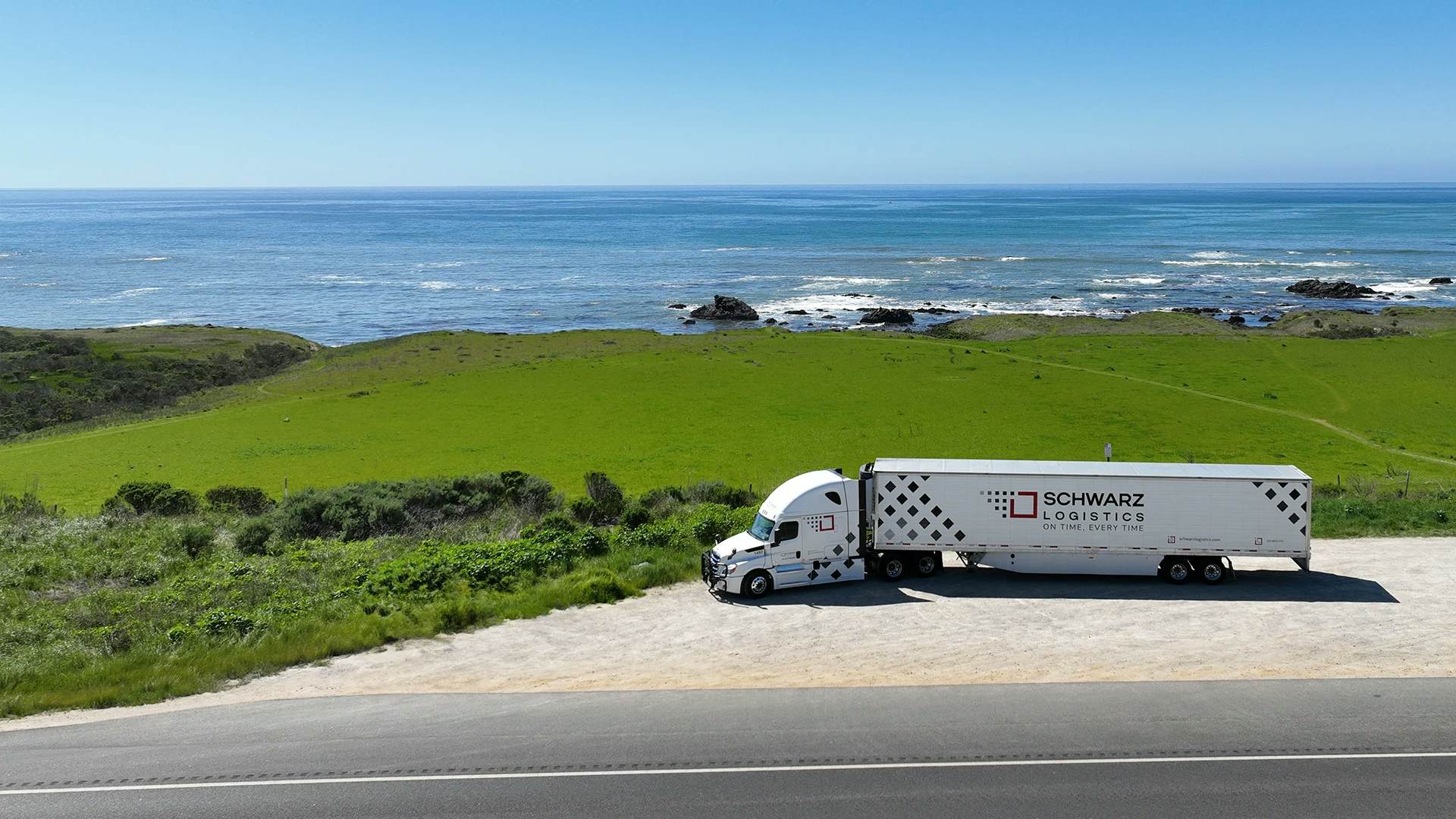 A semi-truck with a white tractor and a long trailer emblazoned with "SCHWARZ LOGISTICS" on the side, suggesting it is part of a logistics and transport company. The truck is parked on a roadside area with a lush green landscape and a scenic view of the sea.